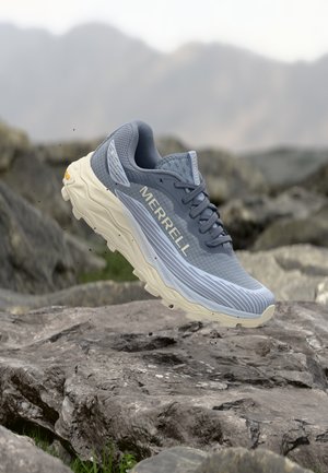 Hellgrauer Trailrunning-Schuh von Merrell schwebt über felsigem Gelände, im Hintergrund sind verschwommene Berge zu sehen.