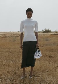 Prigludusi pilka golfą, juoda midi sijonas, sidabro spalvos balerinos, laikanti mažą juodai-baltą dėmėtą rankinę prieš sausų žolių foną.