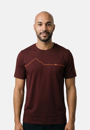 Man met baard en geschoren hoofd draagt een bordeauxrode T-shirt met een eenvoudig oranje berg- en berenlijnontwerp, staand tegen een effen lichte achtergrond.