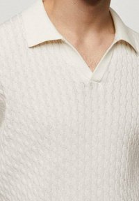 Béžové pletené polo tričko s texturovaným vzorem copánků a měkkým složeným límcem. Má design ve tvaru V výstřihu, který zvýrazňuje detaily látky.