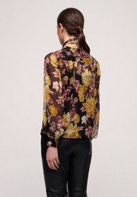 Blusa floreale in tessuto trasparente nero, con grandi fiori gialli e rosa, maniche lunghe e colletto. La vista posteriore mostra dettagli arricciati.