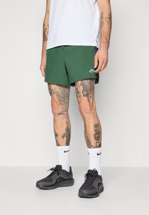 Shorts de sport verts en tissu léger, associés à un haut blanc, des baskets noires et des chaussettes blanches avec un logo. Tatouages visibles sur les jambes.