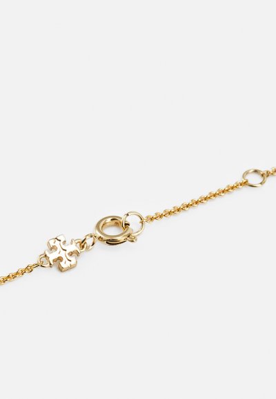 Pulseira de corrente de ouro com um fecho redondo e um pequeno charm em forma de peça de puzzle detalhada, sobre um fundo branco.
