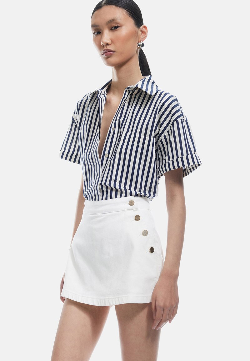 Camicia di cotone a righe in blu navy e bianco con maniche corte, abbinata a una gonna bianca con bottoni decorativi dorati sul lato.