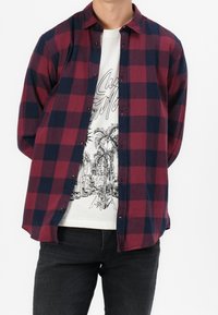 Rot- und marineblau karierte Hemd mit Knöpfen, kombiniert mit einem weißen T-Shirt mit grafischem Druck von Palmen, getragen mit dunklen Jeans.