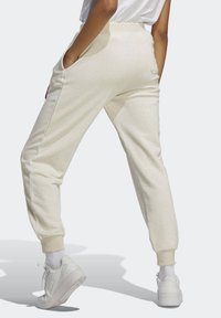 Person som bär off-white träningsbyxor och vita sneakers, står med händerna i fickorna, ett ben något böjt.