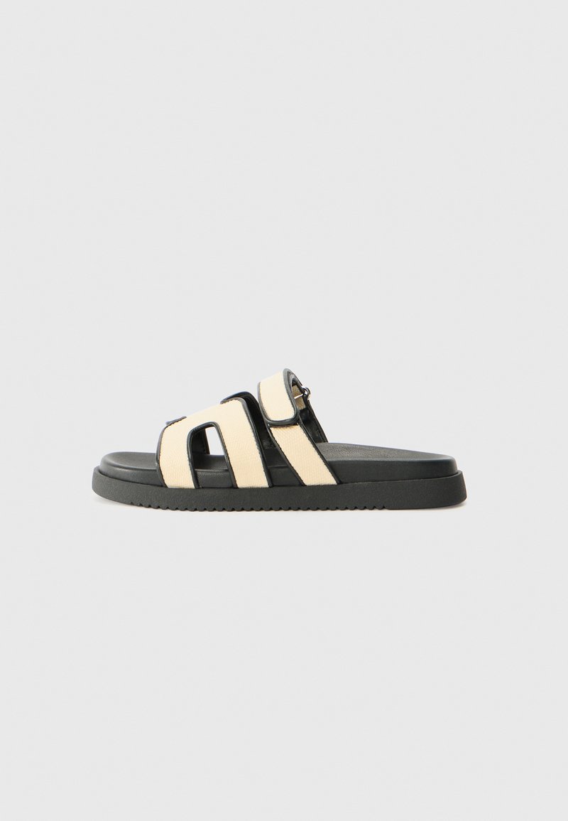 Black and cream slide sandal with two wide straps. Made from synthetic materials, featuring a textured sole and minimalistic design.