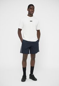 Λευκό κοντομάνικο μπλουζάκι με μαύρο λογότυπο, συνδυασμένο με σκούρο μπλε σορτς με λευκές πλευρικές ρίγες και μαύρα αθλητικά παπούτσια. Μινιμαλιστικός σχεδιασμός.