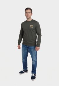 Maglietta a maniche lunghe verde oliva con stampa grafica sulla tasca, abbinata a jeans di denim blu e scarpe scure con dettagli bianchi.
