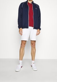 Navy zip-up jacket with white piping, red t-shirt, white shorts, and white sneakers with red accents. Smooth fabric, athletic fit.