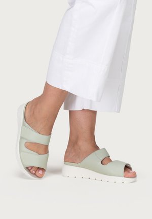 Füße in blassgrünen Sandalen mit offenen Zehen, zwei breiten Riemen und weißen Sohlen, kombiniert mit weißen kurzen Hosen.