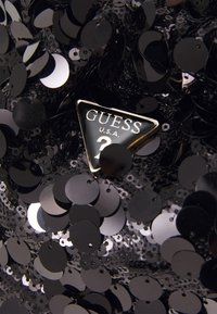 Tessuto nero di paillettes con una texture lucida; presenta un'etichetta metallica triangolare con il logo "GUESS U.S.A." in tonalità bianche e argentate.