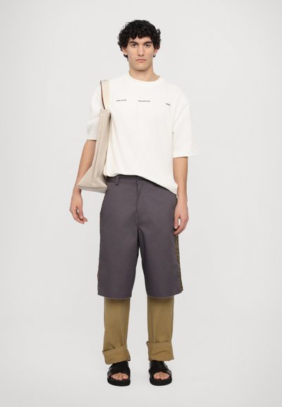 White oversized T-shirt with black text; dark gray shorts with leopard print sides; beige trousers; black sandals; carrying a beige tote bag.