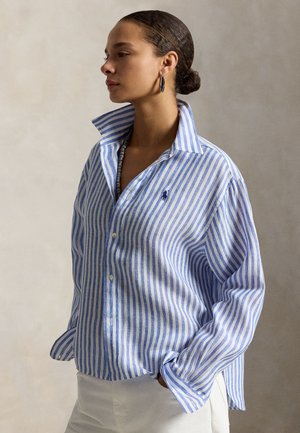 Sieviete valkā zilas un baltas vertikāli svītrainas pogām aizvērtu kreklu ar paceltu apkakli un baltas bikses, stāvot vienkrāsainas fona priekšā.