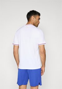 Camiseta atlética blanca de manga corta, con suaves rayas verticales y acentos de color en el cuello y las mangas, combinada con pantalones cortos azules.