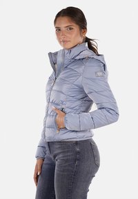 Giubbotto imbottito blu chiaro con zip frontale, cappuccio regolabile, tasche laterali e texture trapuntata. Presenta un'etichetta con logo sulla spalla.