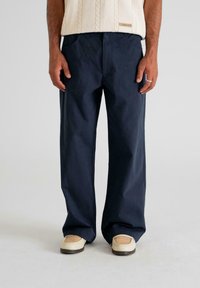 Man wearing navy blue wide-leg trousers, cream knit sweater, beige woven shoes, standing against plain light background.