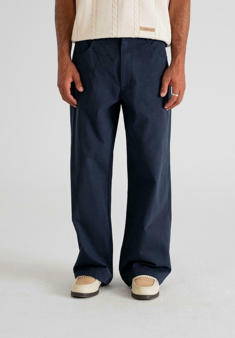 Man wearing navy blue wide-leg trousers, cream knit sweater, beige woven shoes, standing against plain light background.