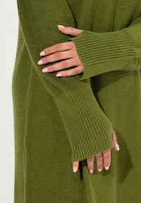 Pull en tricot vert avec des poignets côtelés et une texture douce. Le design comprend des manches longues et une coupe décontractée.