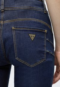 Gros plan sur la poche arrière d'un jean en denim bleu foncé avec une couture jaune et un patch logo Guess triangulaire.