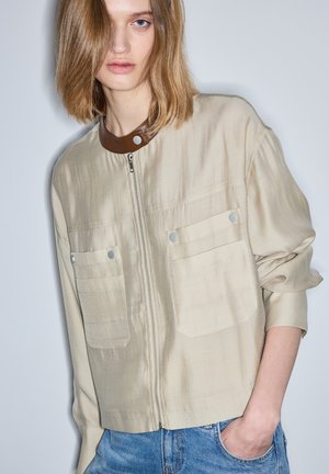 Jonge vrouw met lichtbruin haar tot op de schouders, gekleed in een beige jas met ritssluiting en een bruine kraag, en een blauwe spijkerbroek, hand in de zak.