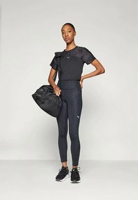 Μαύρο αθλητικό τοπ με κοντά μανίκια, μαύρες κολάν και μαύρα αθλητικά παπούτσια. Μαύρη τσάντα ντουφέκι με λεπτομέρεια από φερμουάρ. Λογότυπο Puma στα ρούχα.