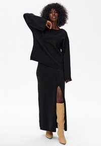 Czarny dziergany sweter o szerokich rękawach w połączeniu z czarną maxi spódnicą z wysokim rozcięciem na udzie, uzupełniony beżowymi kowbojkami sięgającymi kolan.