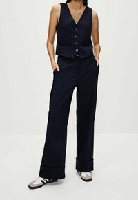 Marineblauw vest met een V-hals en vier knopen, gecombineerd met losse marineblauwe broek met brede omslagen en zakken, aangevuld met sneakers.