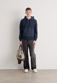 Navy zip-up hoodie with front pocket, paired with dark jeans and white sneakers. Gray and brown tote bag held in hand. Minimalist design.