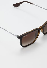 Ruskean kilpikonnakuosiset aurinkolasit, joissa on tummenevat ruskeat linssit, metalliset varret ja näkyvä Ray-Ban-logo kehykseen. Teksturoidut templepäät.