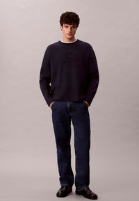 Maglione in pile blu navy con scollo tondo e logo ck in rilievo, abbinato a jeans scuri in denim e scarpe nere. Design semplice, vestibilità casual.