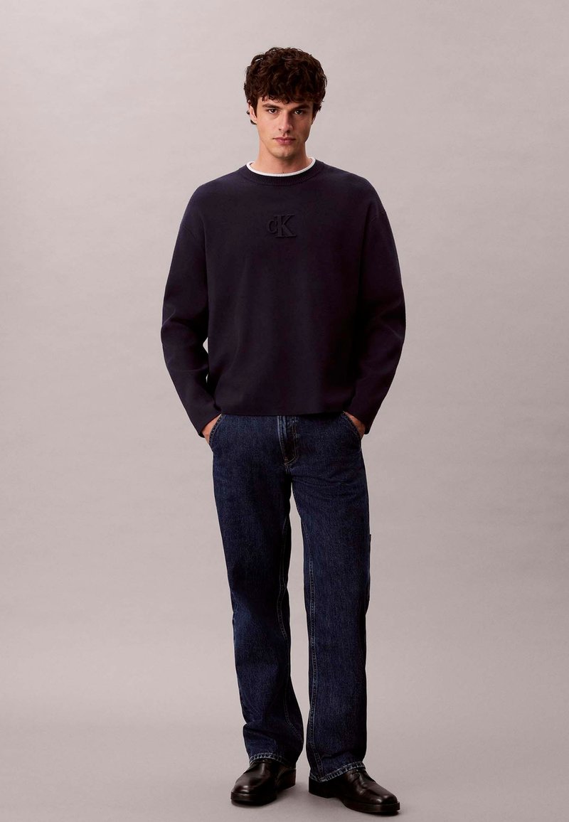 Maglione in pile blu navy con scollo tondo e logo ck in rilievo, abbinato a jeans scuri in denim e scarpe nere. Design semplice, vestibilità casual.