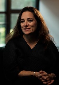 Kobieta z falującymi ciemnymi włosami, ubrana w czarną teksturowaną bluzkę. Siedzi, złożone ręce, na tle z rozmytym tłem z naturalnym światłem.