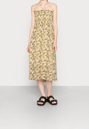 Robe d'été sans bretelles, longueur genou, avec un motif abstrait jaune et gris. Présente un haut smocké et une jupe fluide. Associée à des sandales noires.