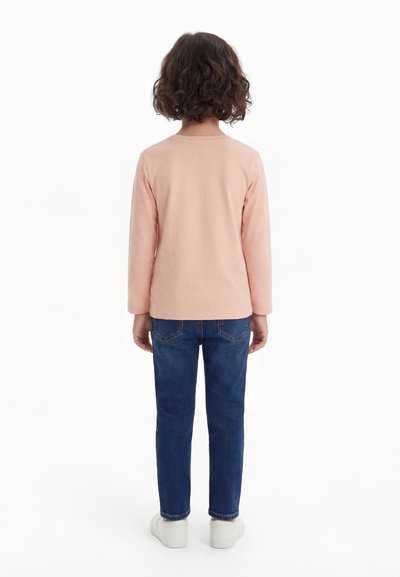 Bērns ar cirtainiem matiem, valkājot gaiši rozā garām piedurknēm krekls, zilas džinsas un baltas kurpes, stāvot ar muguru pret balto fonu.