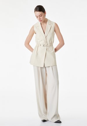 Vrouw met mouwloze crème blazer met ceintuur en crème wijde broek, staand met handen achter de rug.