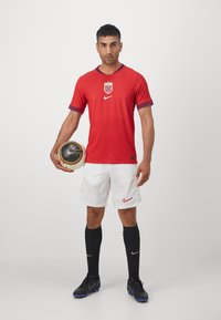 Rød Nike fodboldtrøje med strukturerede striber, der præsenterer det norske flag og logo. Kombineret med hvide shorts, sorte strømper og en fodbold.