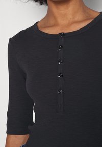 Czarny, dopasowany top z rękawami do połowy przedramienia, o prążkowanej fakturze i okrągłym dekolcie. Posiada sześć czarnych guzików z przodu.