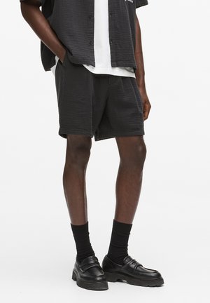 Férfi fekete mintás rövidnadrágban, fekete vastagtalpú mokaszinban, fekete zokniban, fehér pólóban és nyitott fekete rövid ujjú ingben.