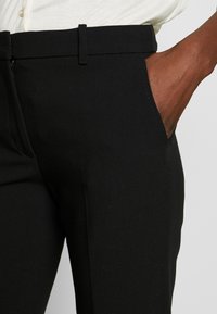 Zwarte broek van gladde stof, met een getailleerde pasvorm, zijzakken en riemlussen aan de tailleband voor een veilige pasvorm.