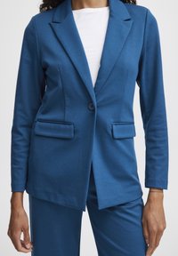Blauer Blazer aus glattem Stoff, mit einem Knopf, zwei vorderen Taschen und einer taillierten Passform. Über einem weißen Hemd getragen.