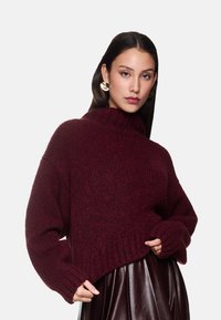 Bordeaux ribbeltrui met een hoge hals en verlaagde schouders, gecombineerd met een glanzende bruine geplooide rok.