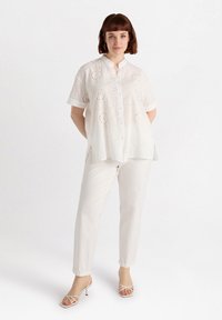 Camicia bianca in cotone con maniche corte e colletto, abbinata a pantaloni bianchi e sandali con tacco alto bianchi.