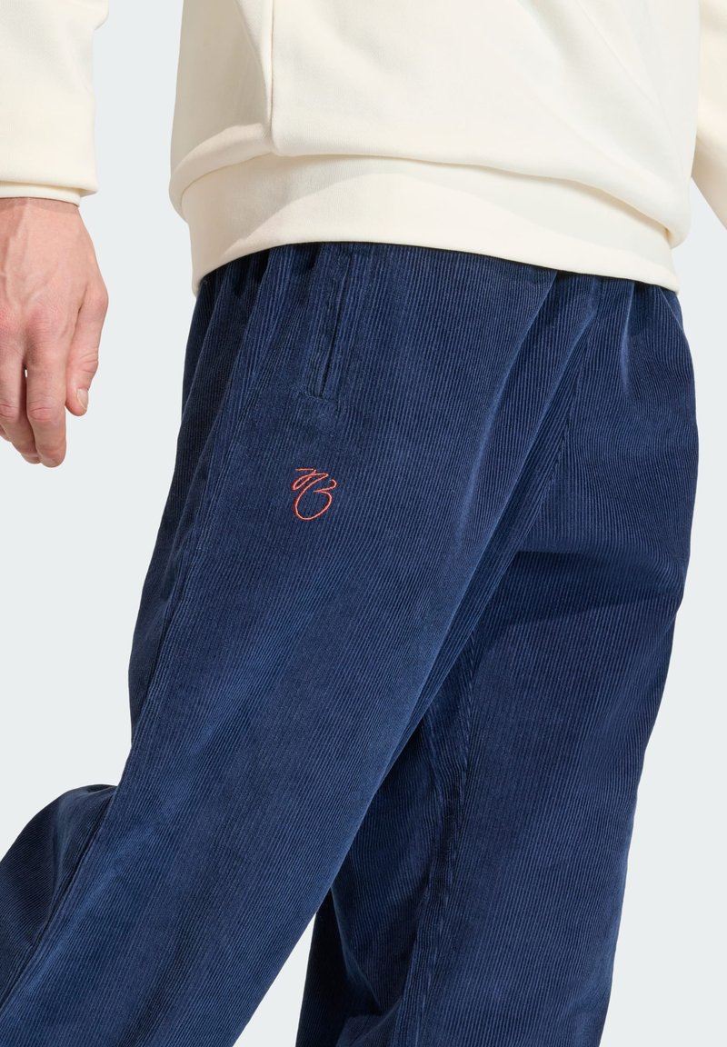 Navy corduroy pants with a relaxed fit, featuring a subtle red logo on the left thigh and side pockets. Textured ribbed surface.