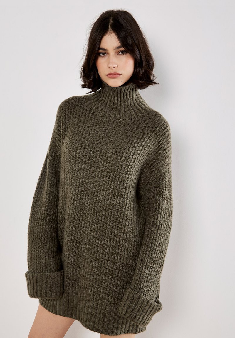 Pull en maille torsadée vert khaki avec un col montant, coupe oversize et poignets larges. Le tissu texturé ajoute de la dimension et de la chaleur.
