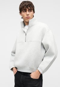 Ανοιχτό γκρι φούτερ με φερμουάρ και ψηλή λαιμόκοψη, με μαλακή υφή και άνετη, φαρδιά γραμμή. Συνδυάζεται με μαύρο τζιν παντελόνι με πλαϊνές τσέπες.