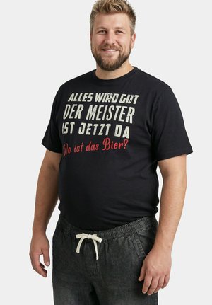 Lächelnder bärtiger Mann in schwarzem T-Shirt mit deutschem Text "Alles wird gut, der Meister ist jetzt da. Wo ist das Bier?" und dunkler Hose mit weißem Kordelzug.