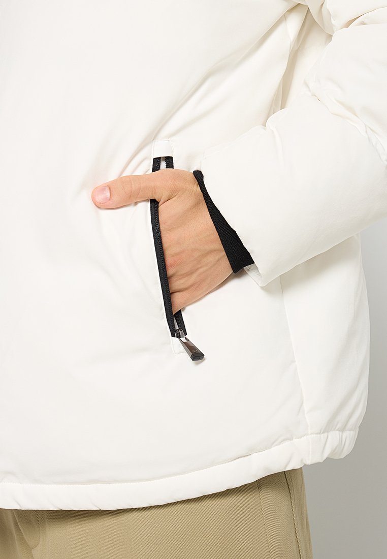Giacca imbottita bianca con una tasca con zip nera. Il tessuto è liscio con polsini elasticizzati, e l'orlo è leggermente elasticizzato.