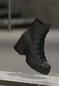 Stivale alla caviglia nero con lacci e tacco largo, realizzato in materiale sintetico testurizzato e dotato di punta rinforzata, posto su uno sfondo neutro.