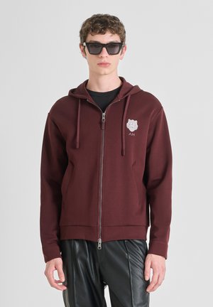 Sudadera con cremallera color marrón en la parte delantera, con un bolsillo, cordones y un logo bordado. Lleva puesta sobre una camiseta negra y combinada con pantalones de cuero negros.